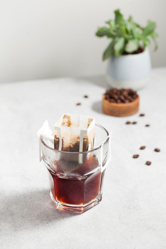 Un filtre Drip Coffee Bag posé sur un verre de café, avec des grains de café et une plante en arrière-plan
